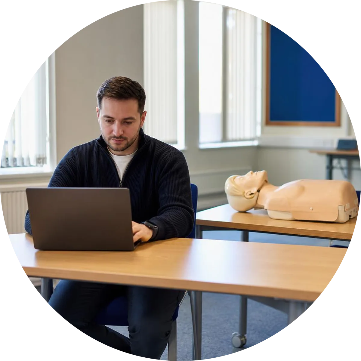 A learner studying online beside a CPR training manikin and notebook